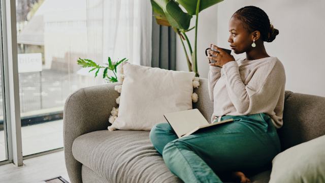 Thoughtful woman in green trousers drinking tea and sitting on a grey sofa
