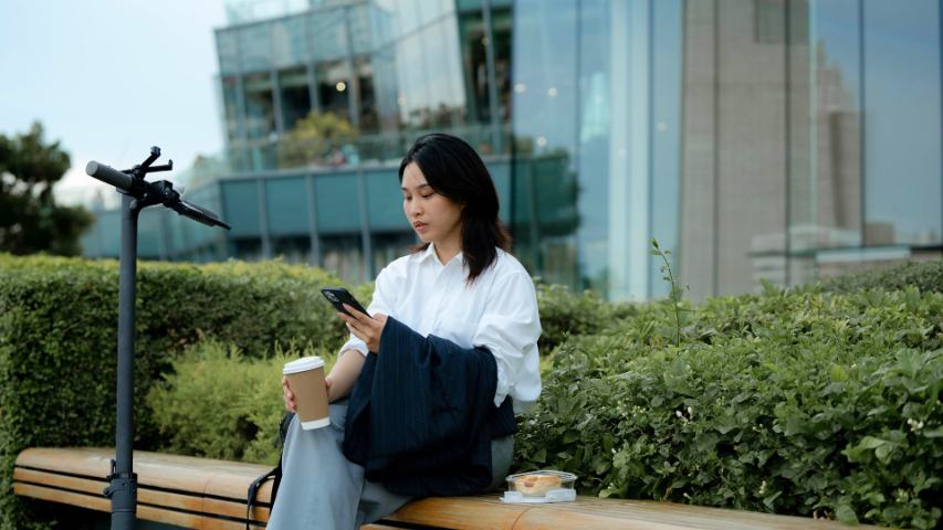 Female sitting on bench outside office building with food and coffee while looking at phone