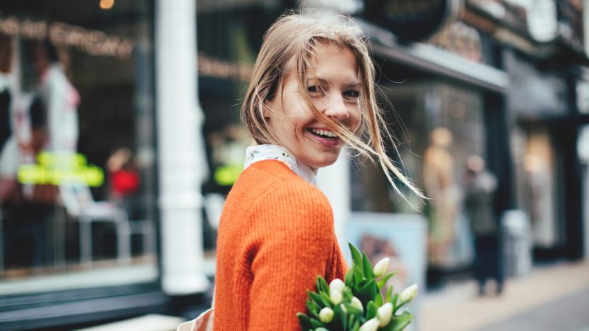 a happy woman with an orange cardigan and tulips smiling over her shoulder as she walks down a street