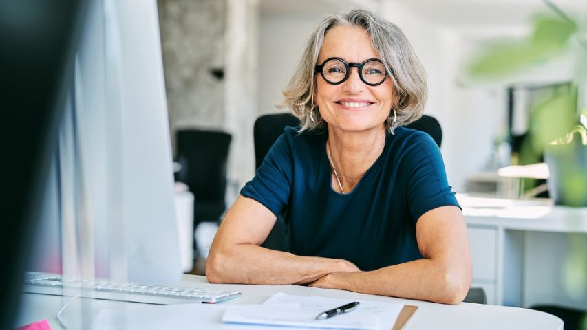 woman with distinctive glasses in a navy tee shirt is smiling directly at the camera while seated at her desk