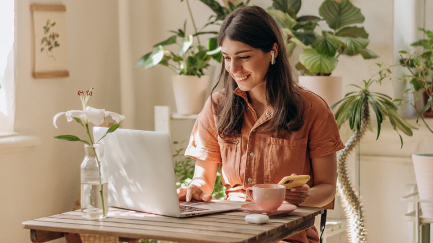 a woman in a shirt dress at a plant filled cafe is using her laptop with earbuds while holding her phone