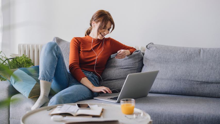 a happy woman using her laptop and credit card while seated on a grey sofa at home