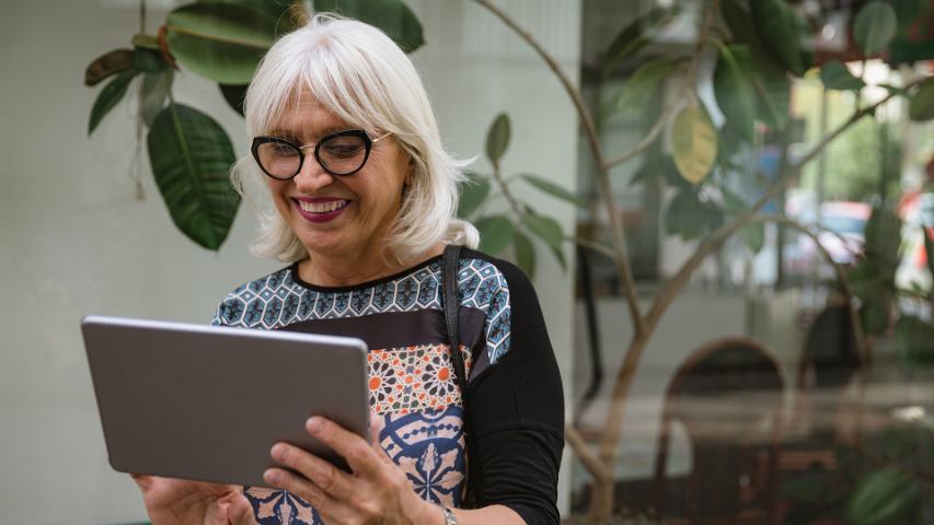 mature woman is smiling while looking at her tablet outside a cafe with a plant behind her