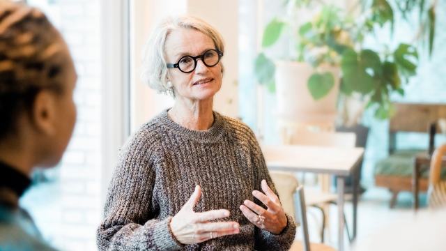 woman in a brown jumper seated at a cafe is describing something  while another woman looks on