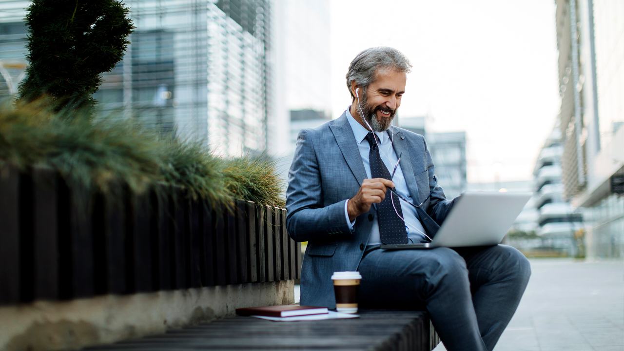 A man in suit and tie doing a meeting outside on his labtop with glasses and coffee 