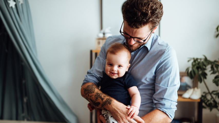 father with glasses in a blue shirt hugs his baby inside their home