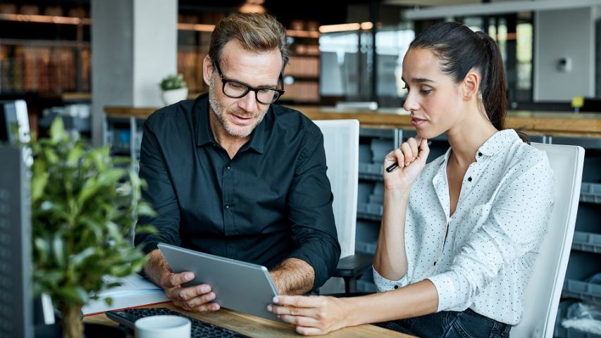 male and female coworker are meeting at a desk and reviewing their work on a tablet in a modern office