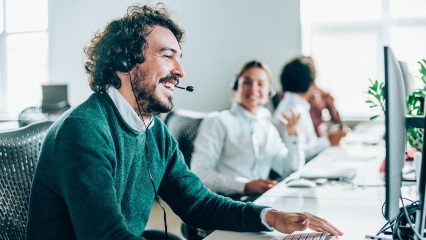 Smiling male wearing headset conducting a call in an office with colleagues in the background