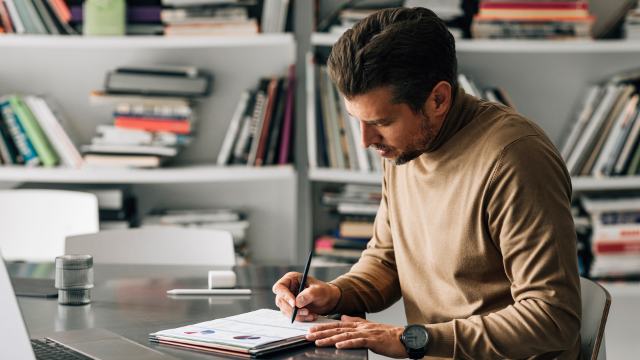 man writing in a notebook while sitting at a deck with books in the background