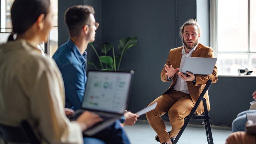 A man in a suit with a tablet presenets to a casual group while they are all seated in an office