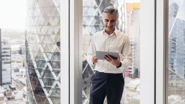 a smiling professional businessman is standing hear the windows of his London office using a tablet 