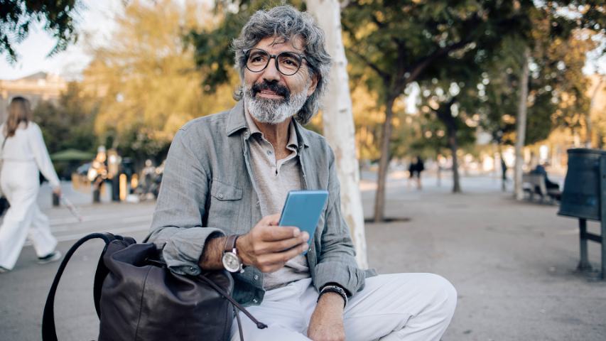 A casually dressed man with a leather backpack is sitting in a park while using his phone and admiring the scenery