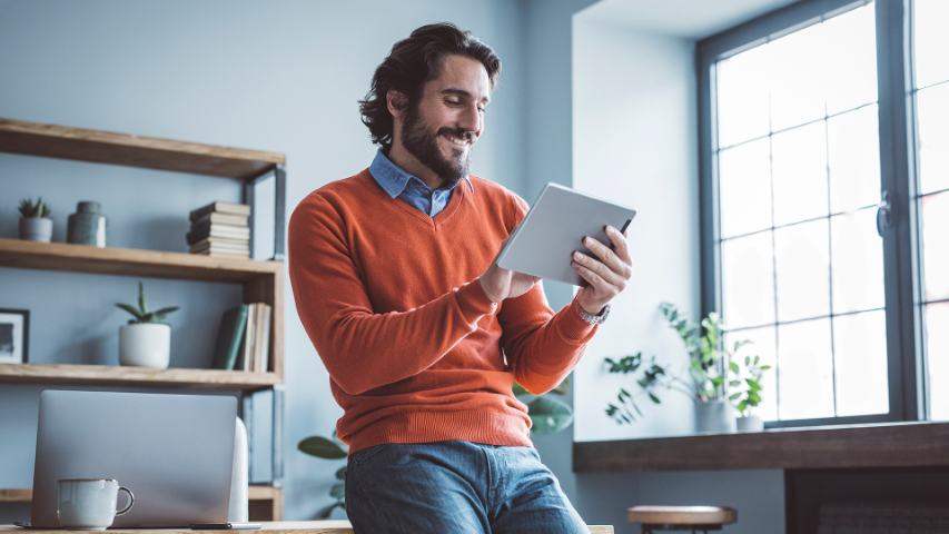 A happy man is his home office is smiling while using his tablet