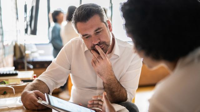 A man and woman are having a casual meetng to discuss a project at her desk and are looking at a tablet