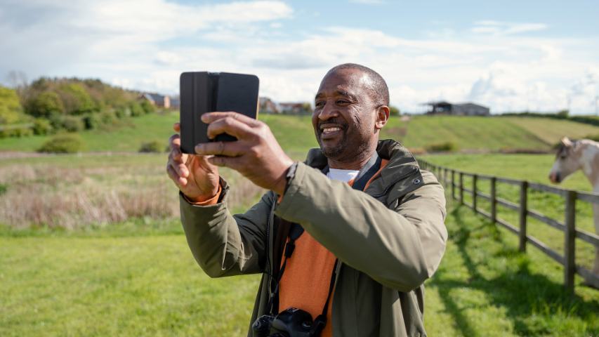 man with a camera around his neck is using his phone while standing in a rural field
