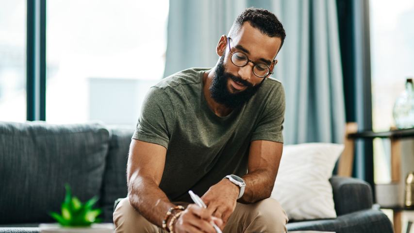 man in glasses and a green tee shirt is writing something down while sitting on his sofa at home