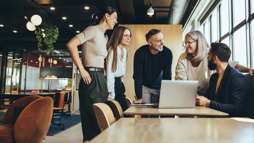 Mixed group of smiling male and female professionals gathered around a laptop in discussion