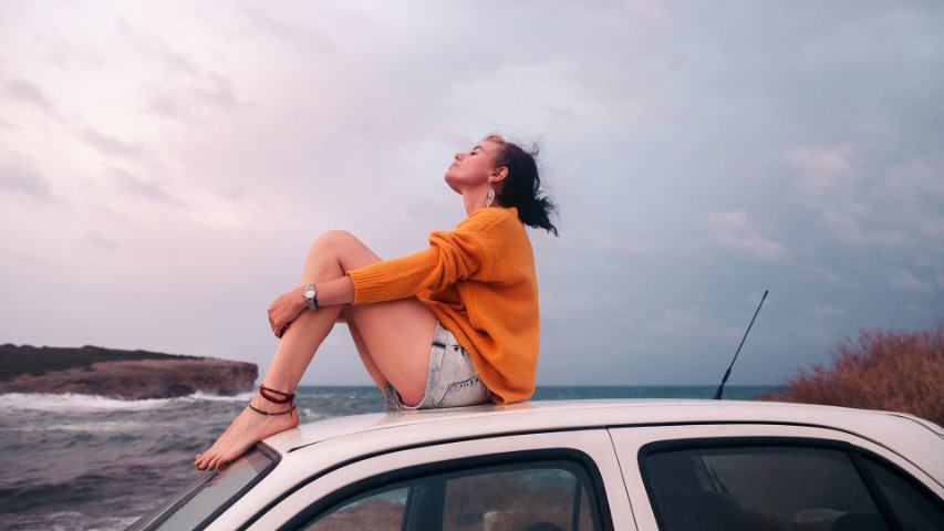 Young woman in an orange jumper sitting on the roof of a cream car by the sea with a cloudy sky