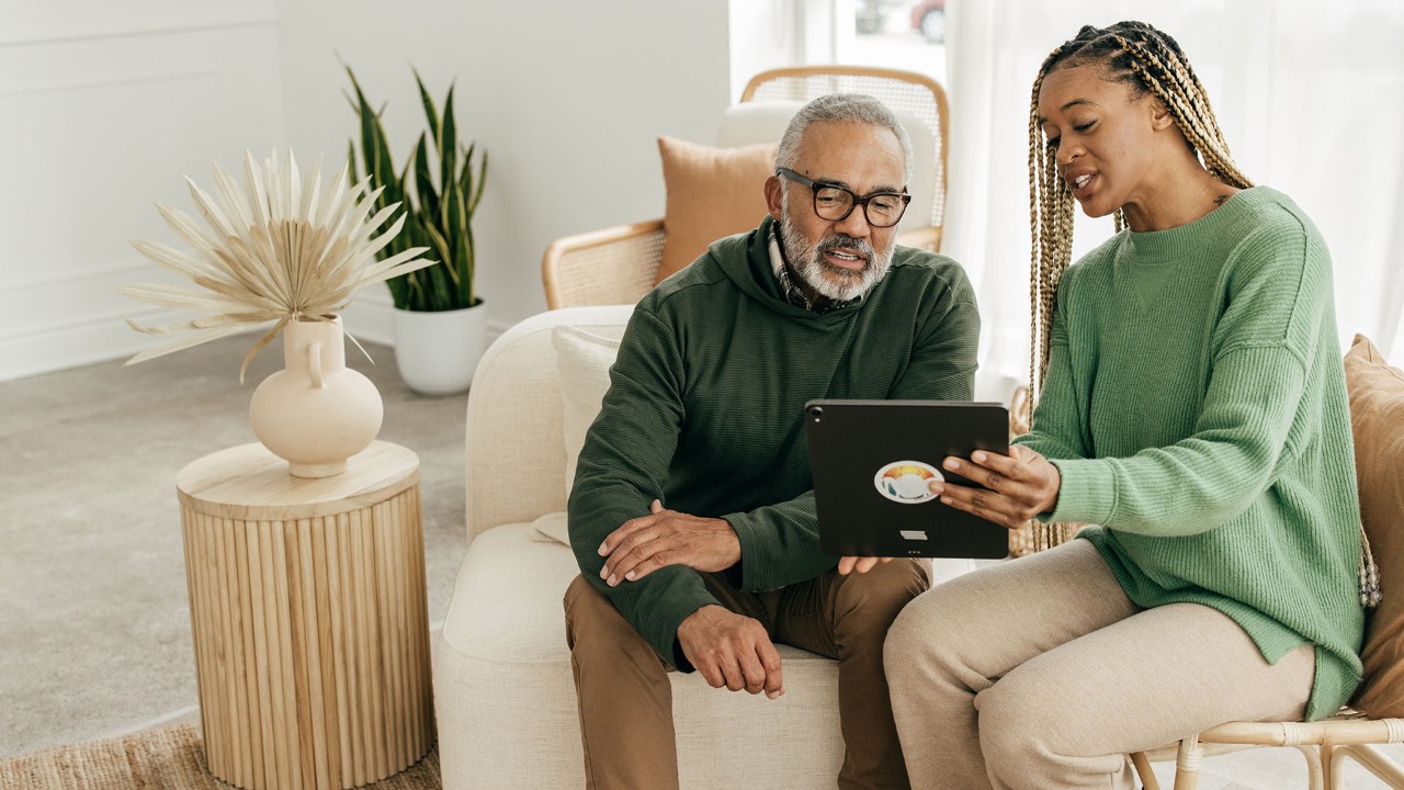 Woman with braids and a green sweatshirt is showing a tablet to an older man as they sit on a cream sofa