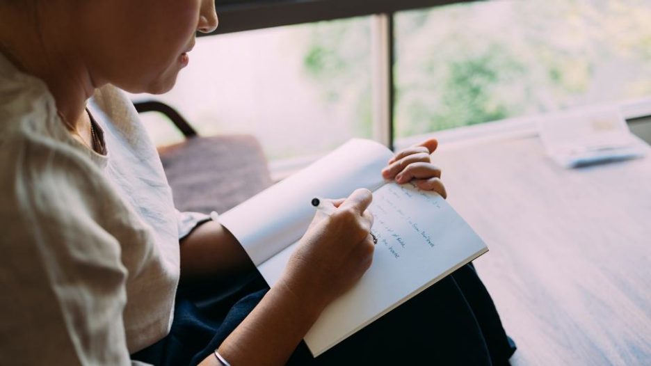 woman writing in a hotebook while sitting near a window at home