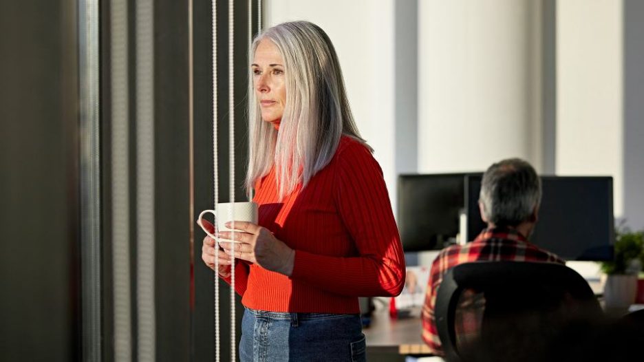 Woman with coffee cup in office looking out window thoughtfully