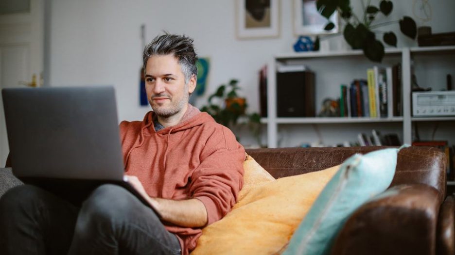 man in a hoodie is using his laptop while sitting on his sofa at home