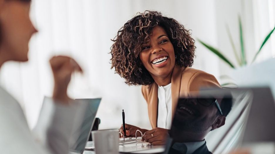 a professional woman  writing in a binder at her desk looks up to smile at something a coworker has said