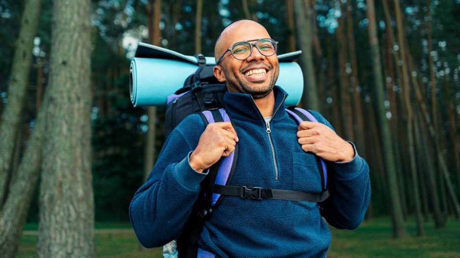 a man in glasses with a hiking bag  on his shoulders smiles walking through the forest on a summer day