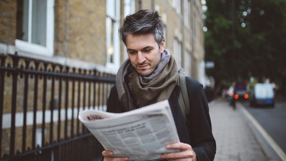 Vintage toned portrait of a young man in London, waiting for the public transportation or just slowly walking home from work, reading the newspapers. He is casually dressed, wearing a denim jacket and a scarf.