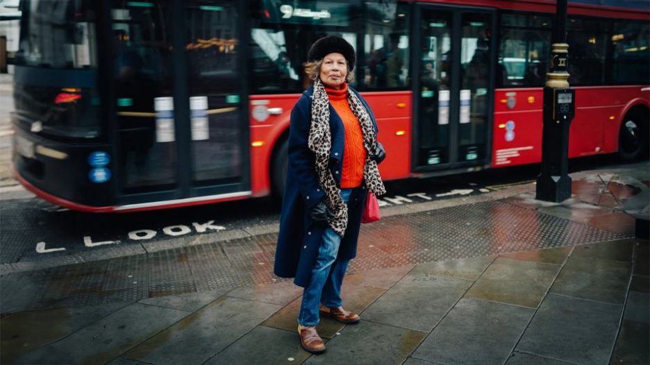 older woman standing on pavement while bus drives behind her.