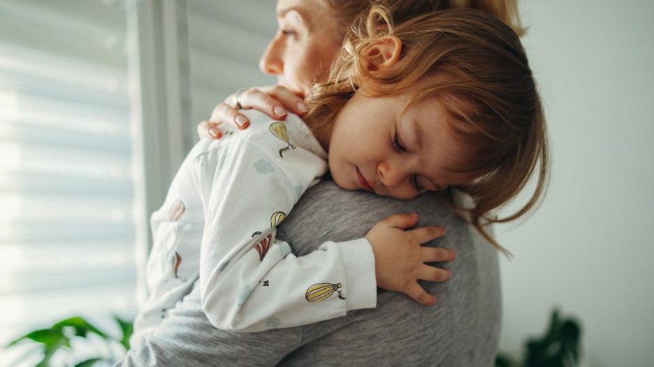 woman with a sleeping toddler on her shoulder looks out a window in the early morning