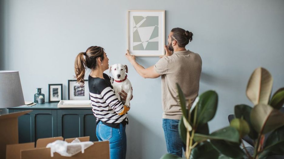 husband is hanging a new picture on the wall while his wife looks on and cuddles their dog