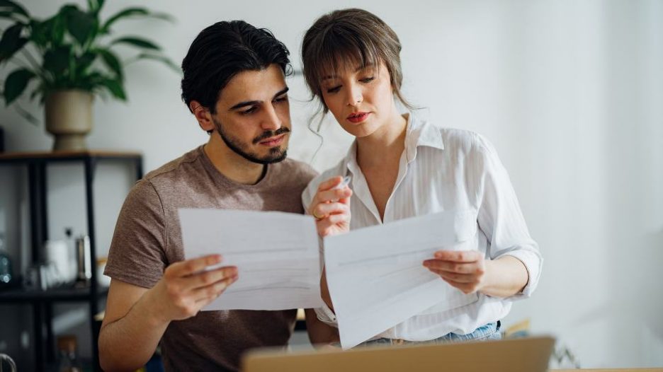 man and woman sitting slose together to compare paperwork with a laptop nearby man and woman sitting slose together to compare paperwork with a laptop nearby
