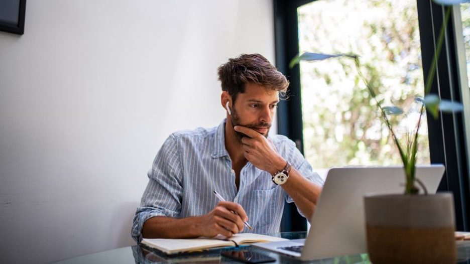 man in a striped shirt using his laptop and taking notes in a home office