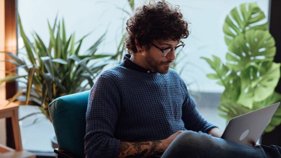 thoughtful man with glasses and a tattooed forearm is working on his laptop in a modern space with plants