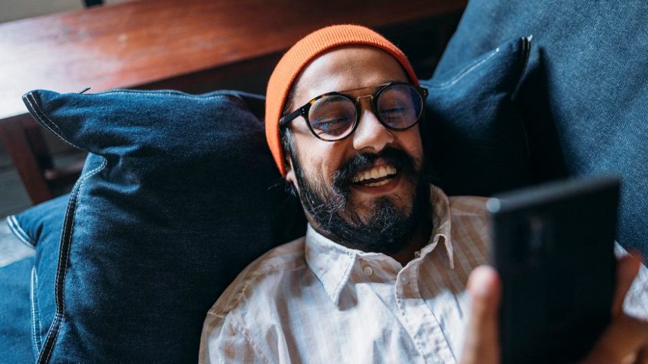 Bearded man in orange hat lying on the couch looking at his phone