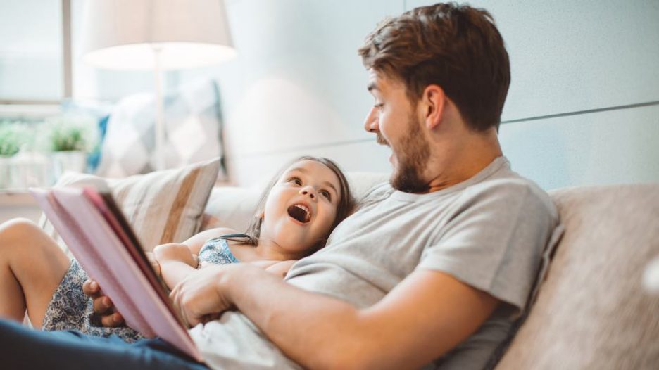 father is reading an exciting book to his daughte ras they lie on a bed