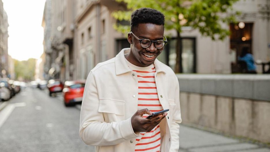 smiling man with glasses in a cream jacket using his phone while walking down a street