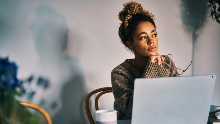 woman in a brown jumper pauses while working on her laptop to think and drink coffee