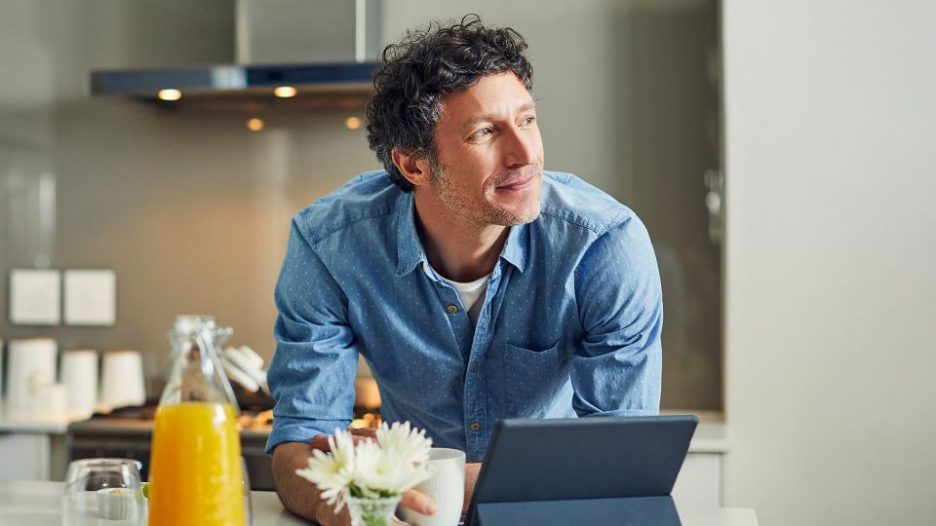 man drinking tea in his kitchen and looking hopeful with his tablet nearby