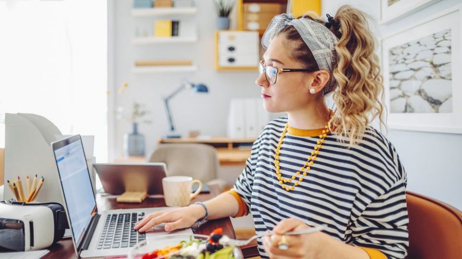 young woman in a striped top is working on a laptop and eating her lunch in an office