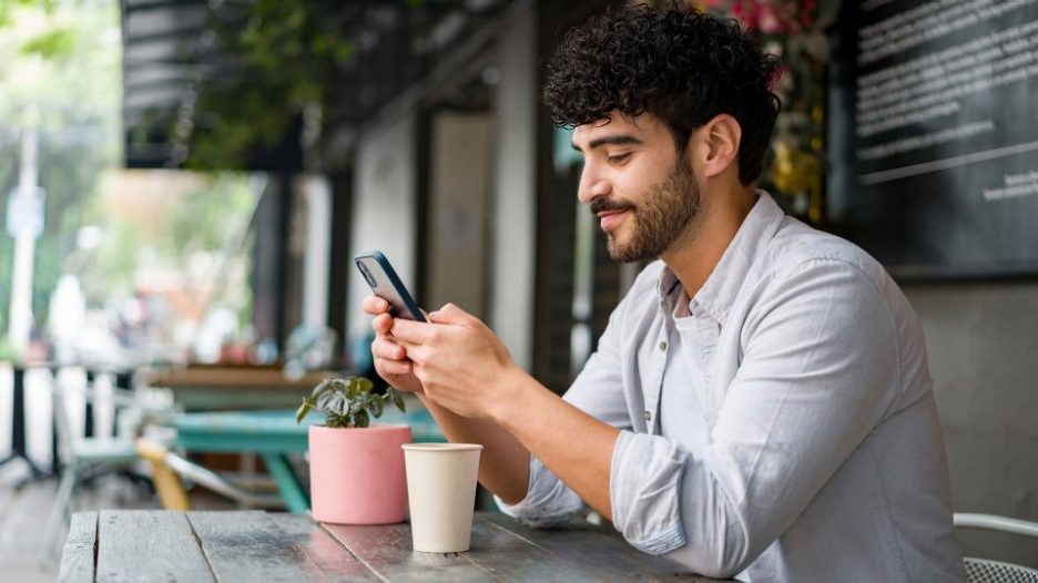 A happy man is using his phone while having a coffee at an outside table of a cafe