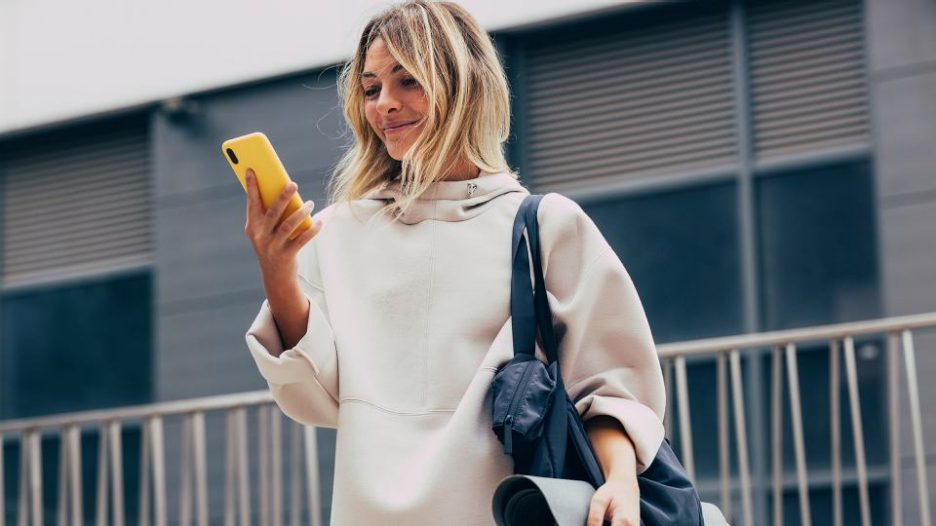 a woman with a sports bag and yoga mat is smiling at her phone while standing outside
