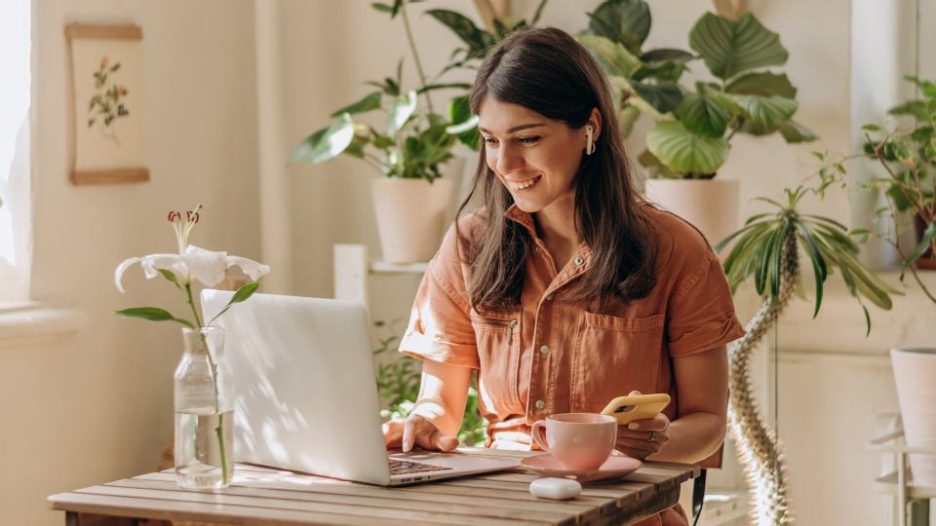 Female smiling using laptop, phone and earbuds at table surrounded by plants