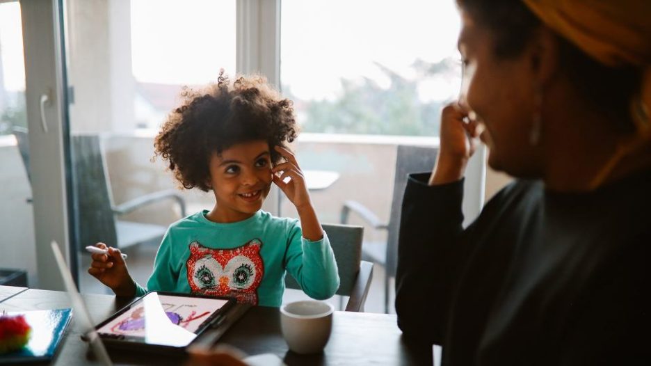 a happy young girl doing art on an ipad at a table inside while a woman looks on