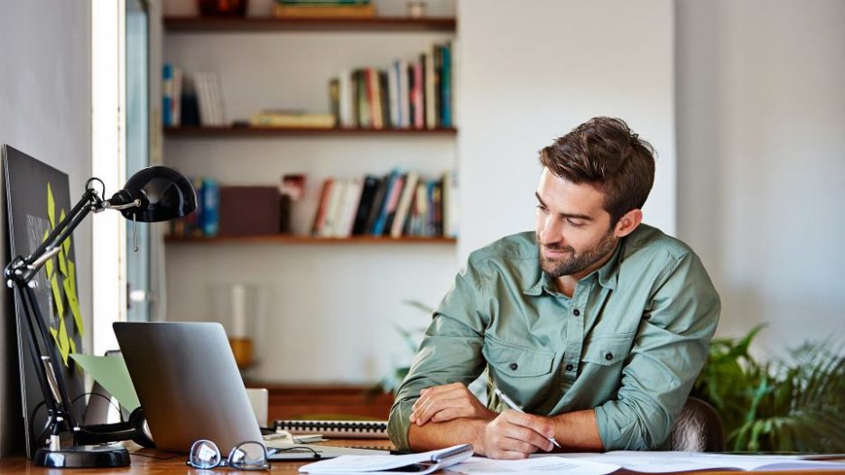 relaxed man in his home office is looking at his laptop and taking notes