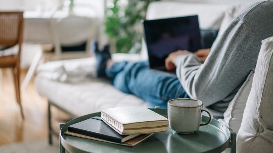 a man whose face we cannot see is lying on his sofa with a laptop and coffee