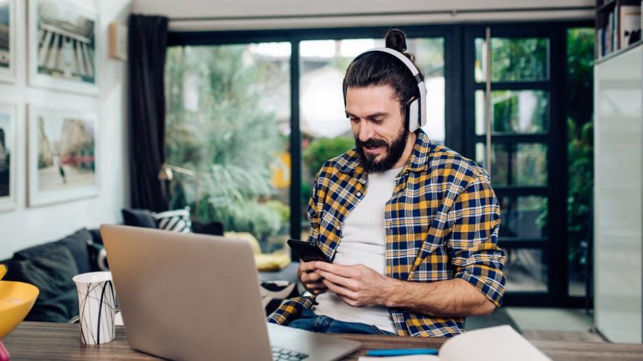 man in a checked shirt with headphones using his phone while seated at home with his laptop nearby