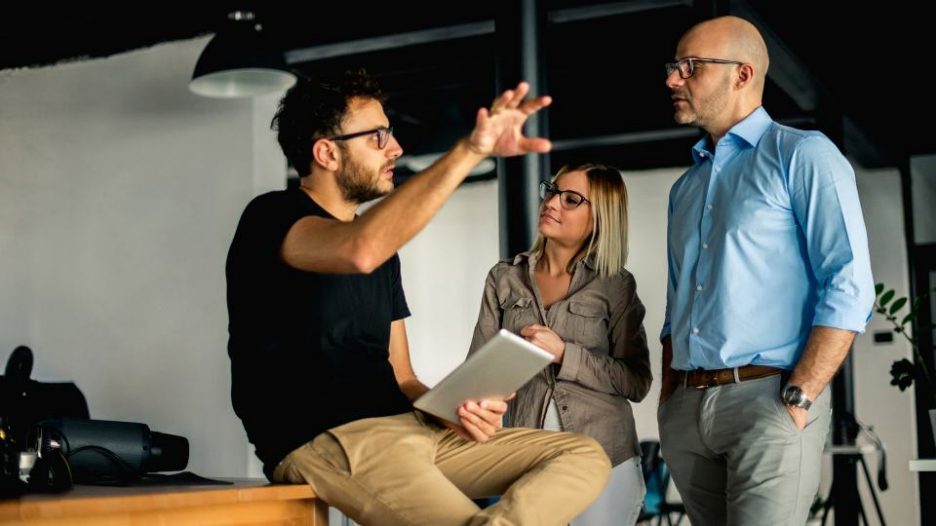 one man describes his ideas to a man and woman while in a relaxed office setting