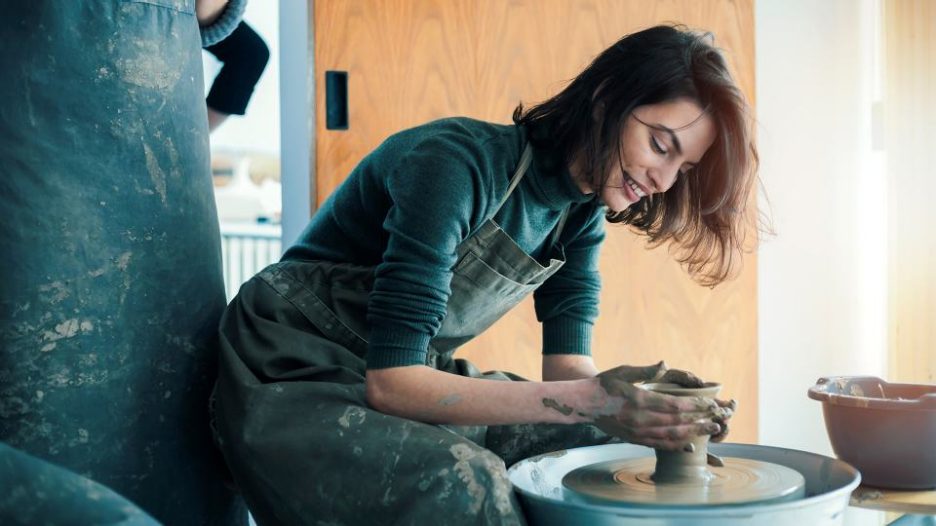 a woman in a green apron turns clay on a wheel while another potter looks on